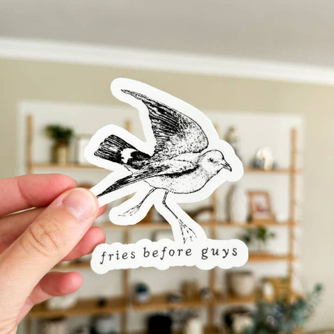 Hand holding a sticker with a bird design and text 'fries before guys' in a room setting.