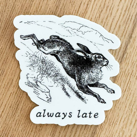 Sticker with a black and white illustration of a rabbit and the text 'always late' on a wooden surface.