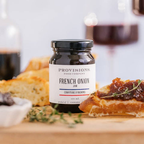 Jar of Provisions French Onion Jam on a wooden board with bread and herbs.
