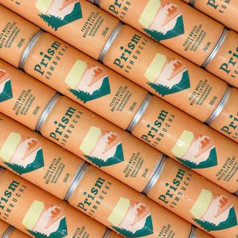 A close-up, repeating view of multiple Prism Kombucha cans arranged diagonally. The cans are a warm peach-orange color with a printed graphic of a mountain and forest, and the brand name 'Prism' is clearly visible.