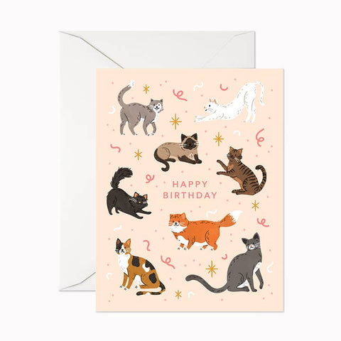 A birthday greeting card with a light peach background, decorated with multiple illustrated cats in different poses and the words 'HAPPY BIRTHDAY' in the center. A white envelope is placed behind the card on a clean white background.