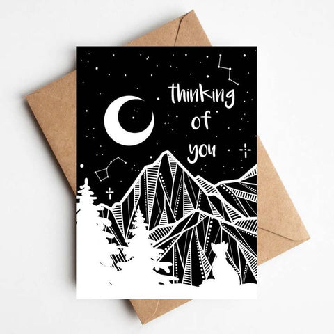 A black greeting card with a white line-art night sky design, including a crescent moon, stars, constellations, mountains, trees, and the phrase 'thinking of you,' shown with a brown kraft envelope on a white background.