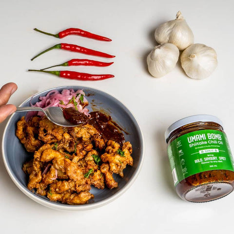 Bowl of fried chicken with a side of sauce, red chilies, and garlic bulbs on a white background.