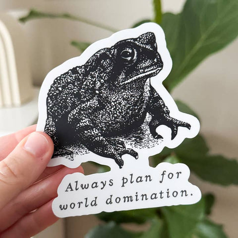 Sticker with a frog design and text 'Always plan for world domination' held by a hand.