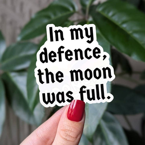 Hand holding a sticker with text 'In my defence, the moon was full' against a leafy background