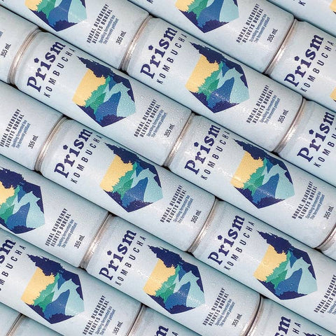 A close-up, repeating view of multiple Prism Kombucha cans arranged diagonally. The cans are a very pale icy blue with a graphic of layered green and blue mountains with a yellow sun, and the brand name 'Prism' and the word 'Kombucha' printed on the front.