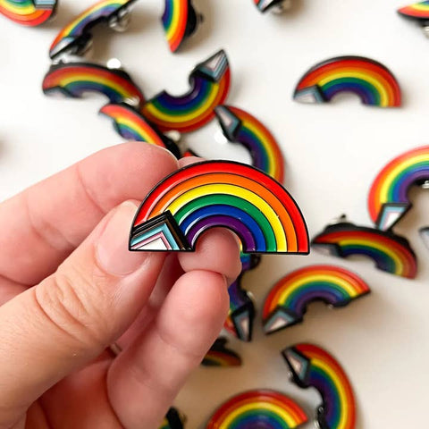 Hand holding a rainbow-shaped pin with more pins in the background on a white surface