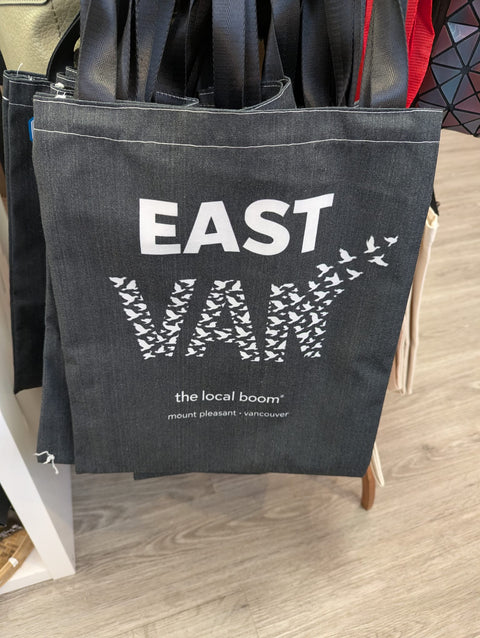 Black tote bag with 'EAST VAN' design and 'the local boom' text on a wooden floor.