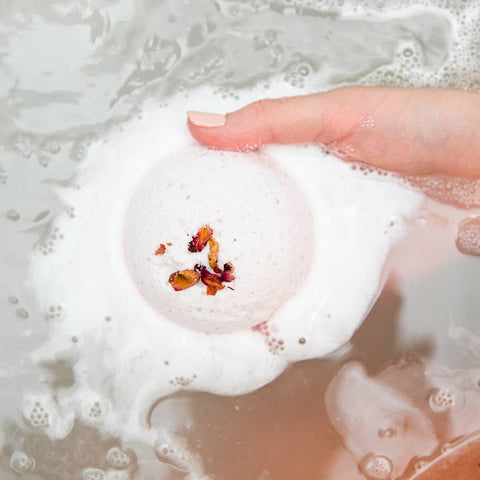Foamy bath bomb with rose petals being held above a bathtub filled with water and bubbles.