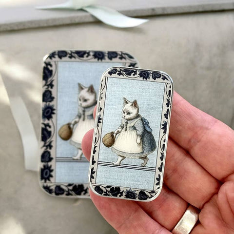 Small rectangular tin with a cat illustration held by a hand, with another similar tin in the background.