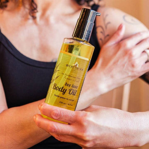 A person holding a clear bottle of bee love body oil with a black pump dispenser, while another person applies the oil to their upper arm in a close-up shot.