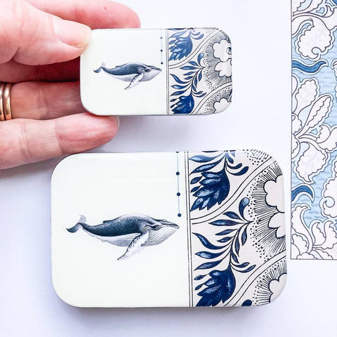 Small decorative tin with whale and floral design held by a hand on a light background