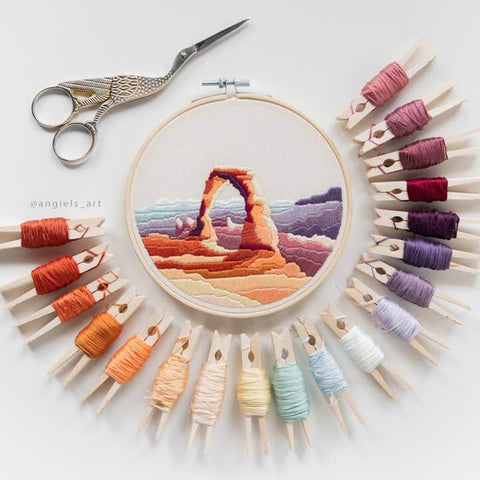 Embroidery hoop with a colorful landscape design surrounded by yarn spools and clothespins on a white background.
