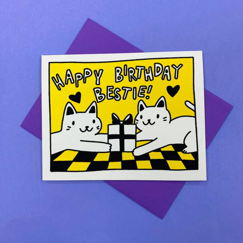 A square greeting card with a white border, featuring a yellow background and cartoon cats around a wrapped gift box, with the text 'HAPPY BIRTHDAY BESTIE!'. The card is placed on top of a purple envelope against a solid purple background.